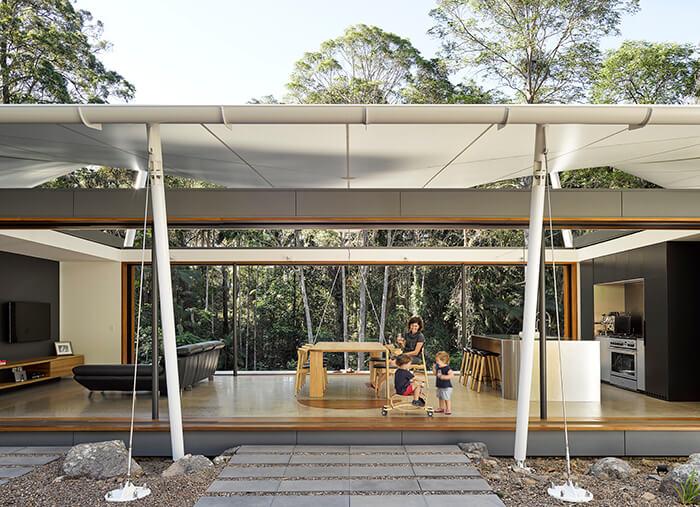 Mãe e dois filhos brincam na área ocupada por sala e cozinha abertas da Tent House. Atrás, paredão da floresta