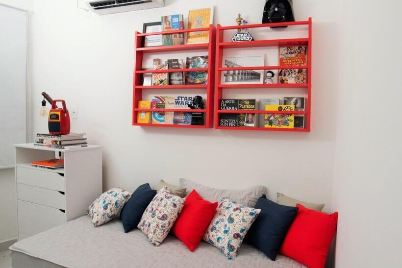 quarto de hóspedes com detalhe para a parede onde a cama está encostada, onde se encontram duas estantes de parede vermelhas penduradas para armazenar livros